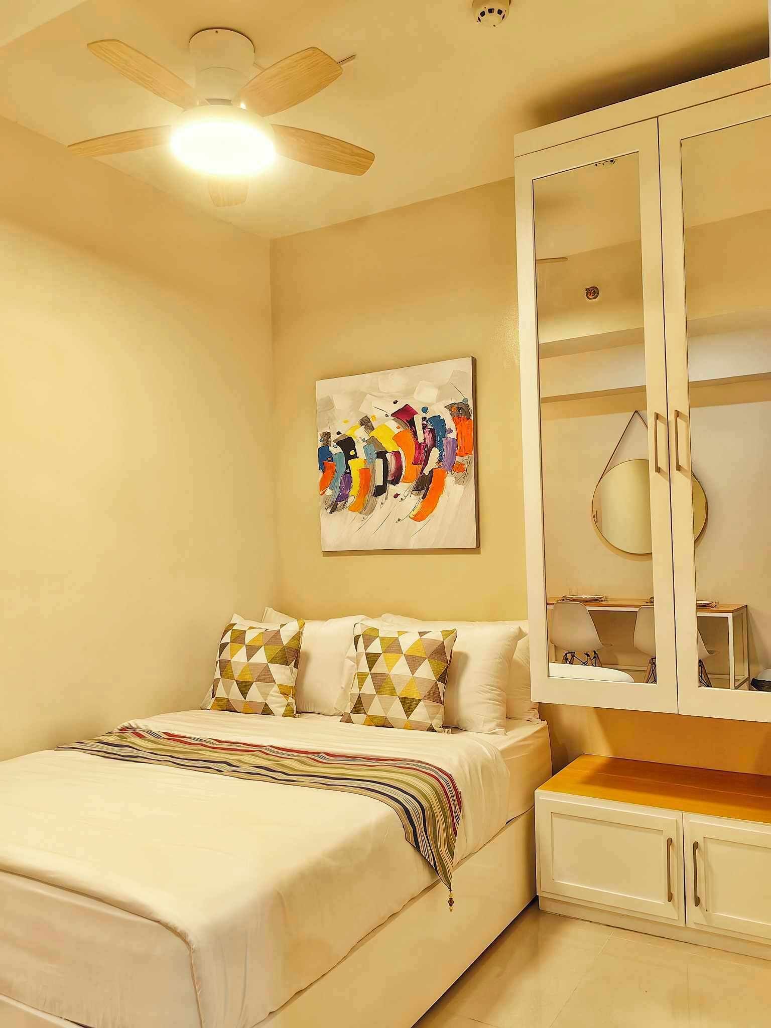 A bedroom with a bed, a ceiling fan, a painting, and a wardrobe.