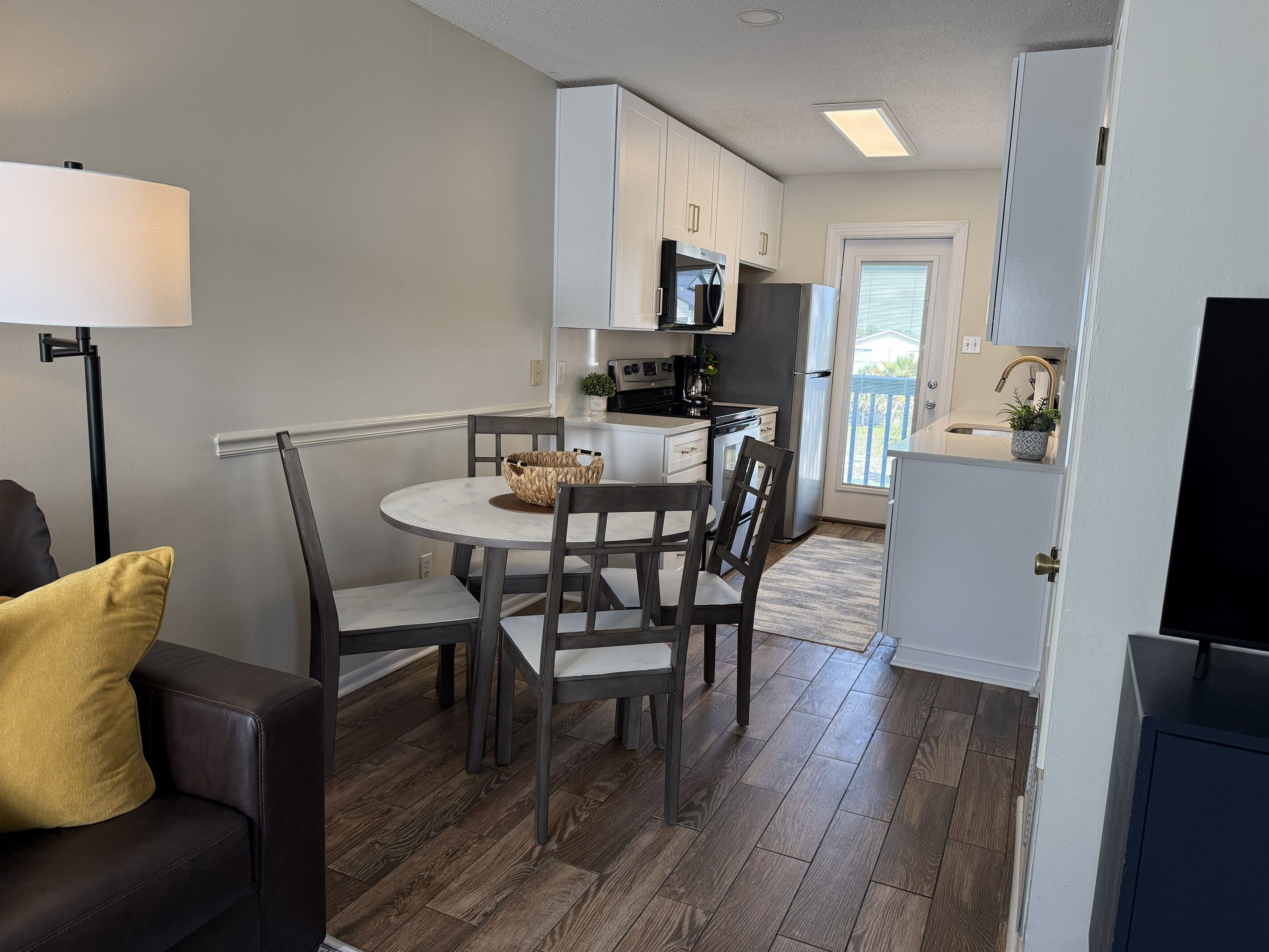 A compact kitchen with white cabinets, a microwave, and a refrigerator. There is a dining table with chairs and a floor lamp.