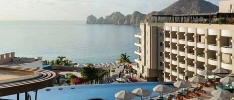 Zona de piscina en un resort con múltiples tumbonas y sombrillas, una piscina grande y vistas al mar y a las montañas.