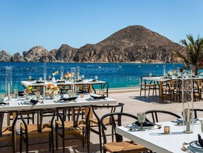 Uma área de refeições ao ar livre com mesas para dois, com vista para um mar azul e límpido e colinas rochosas.