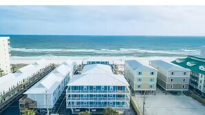 A coastal hotel with a central building featuring a blue facade and multiple windows, surrounded by other buildings and a beach view.