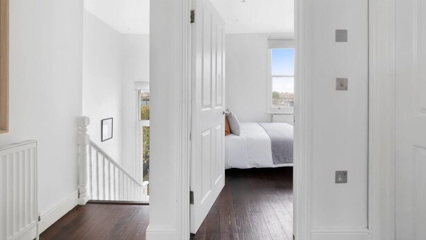 Een gang die leidt naar een slaapkamer met een bed, witte muren en uitzicht op buiten.