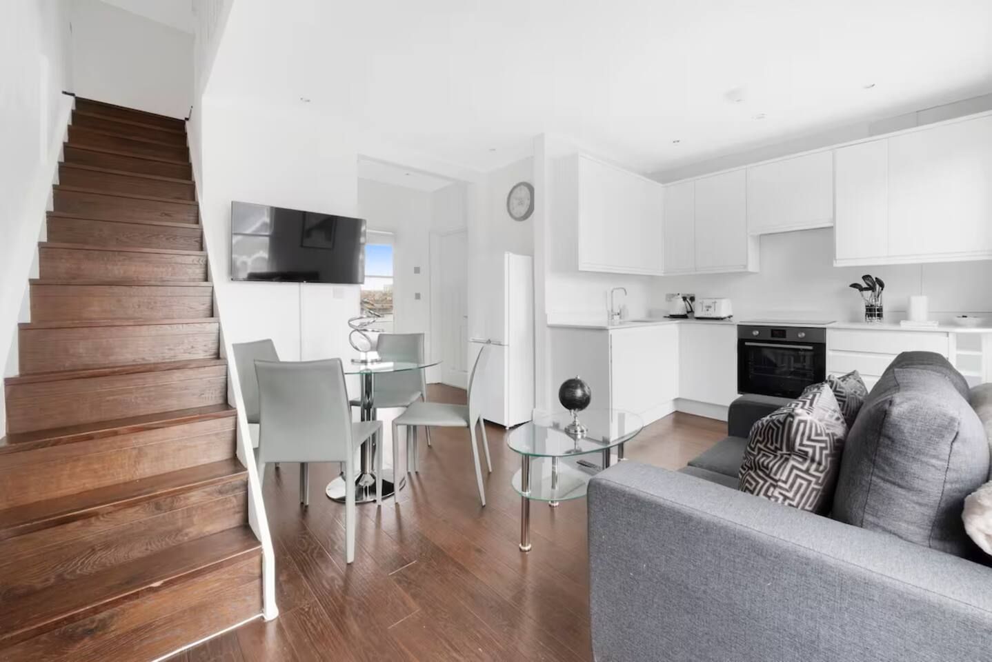A modern apartment with a wooden staircase, a dining area with a glass table, a kitchen with white cabinets, and a grey sofa.