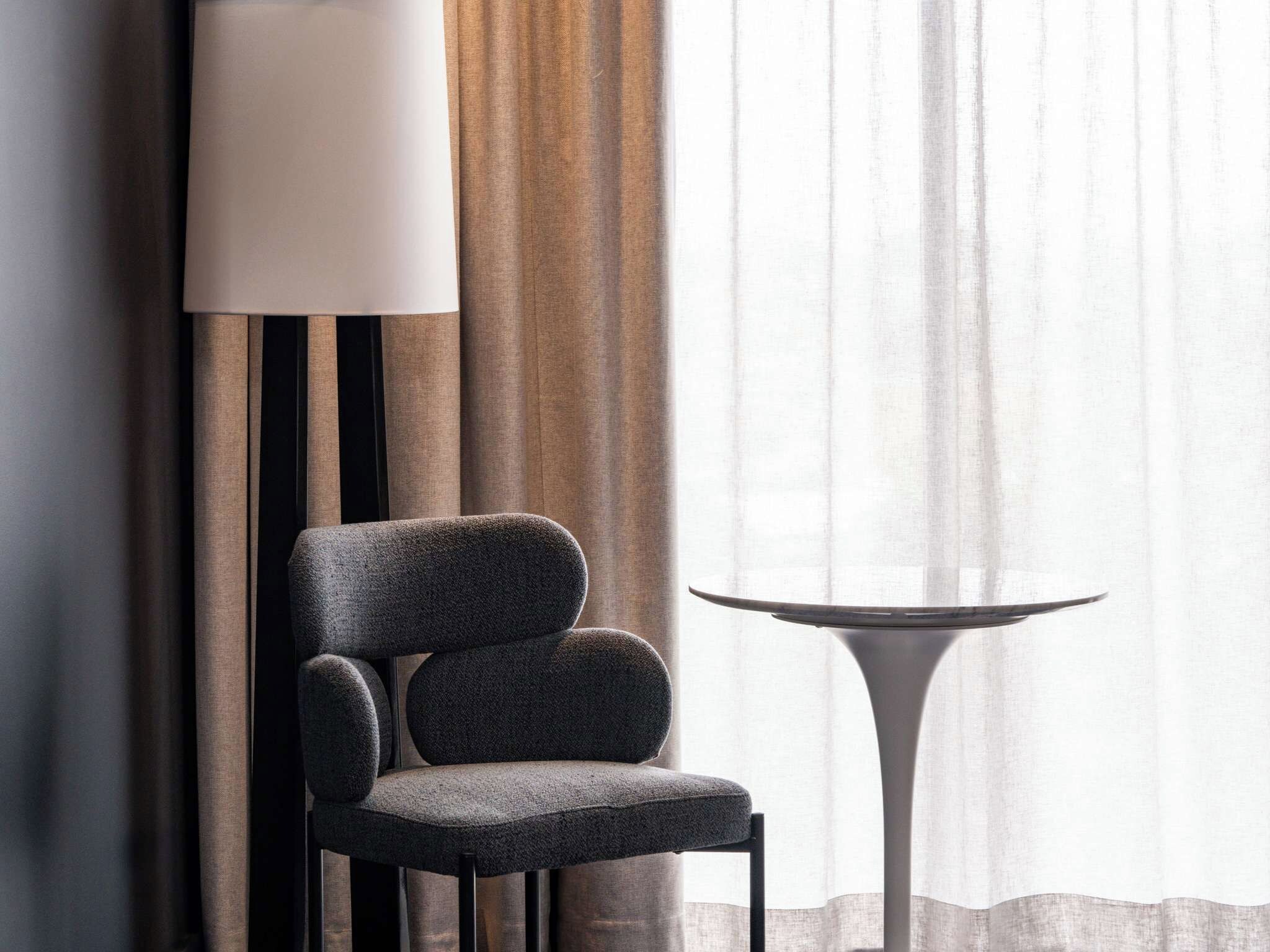 Una silla moderna y una mesita en una habitación con un ventanal, cortinas y una lámpara.