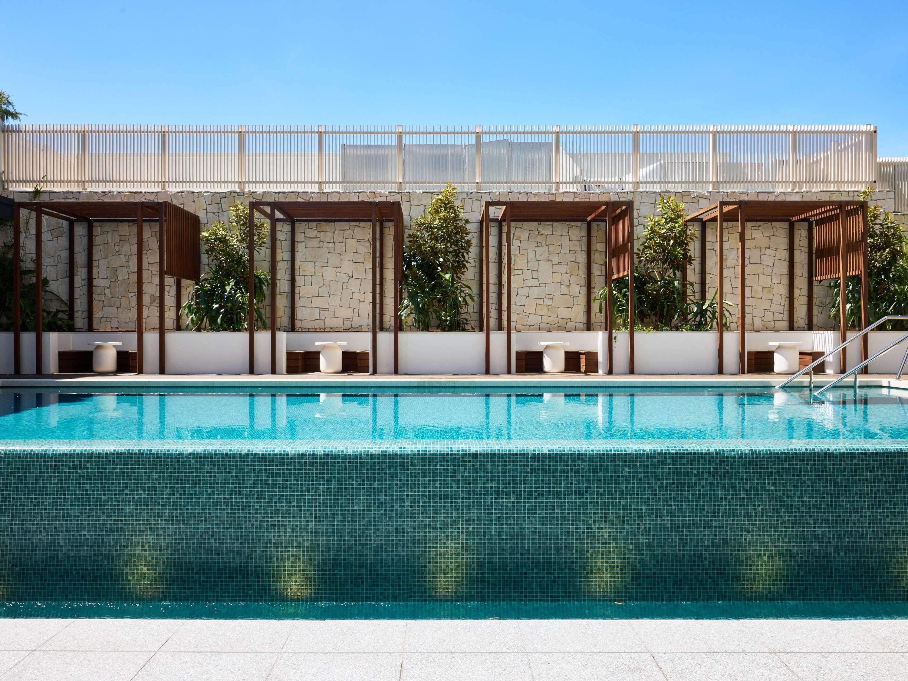 Una piscina con muro de piedra, pérgolas de madera y zonas de estar.