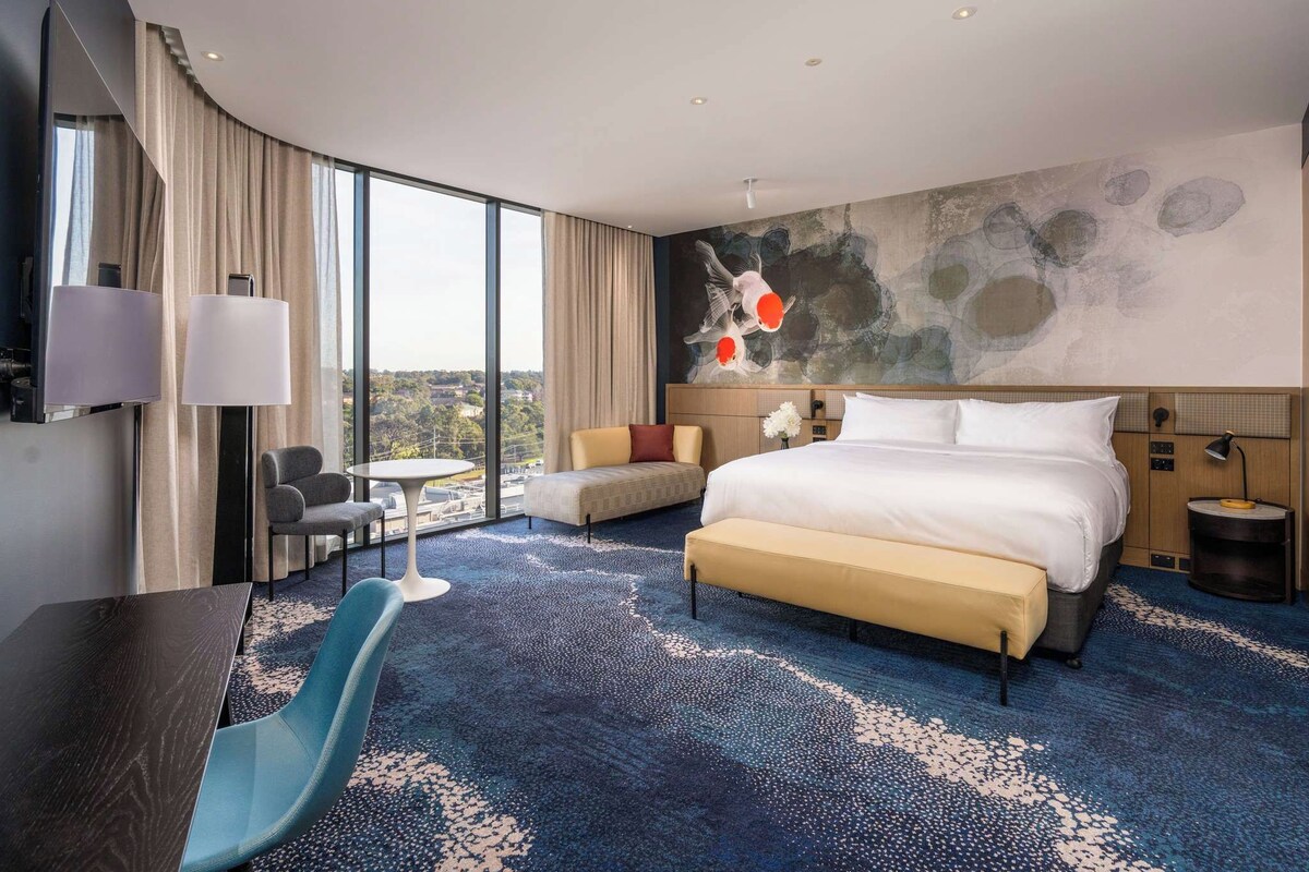 A modern hotel room with a large bed, a desk, a chair, a small table, and a view of the city.