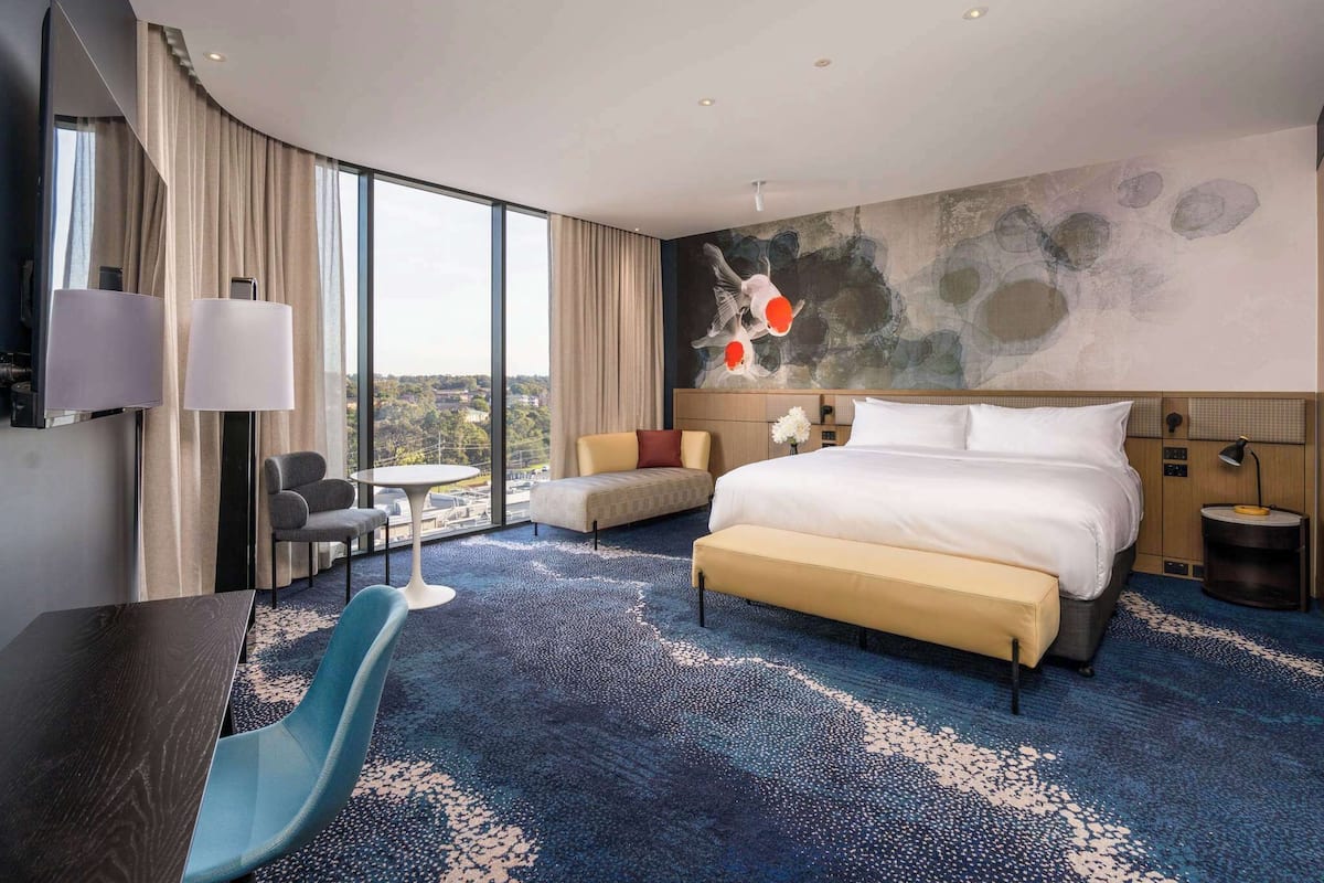 A modern hotel room with a large bed, a desk, a chair, a small table, and a view of the city.