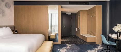 Una habitación de hotel moderna con una cama grande, un banco, un escritorio con una silla y vistas a la zona de ducha a través de un vano.