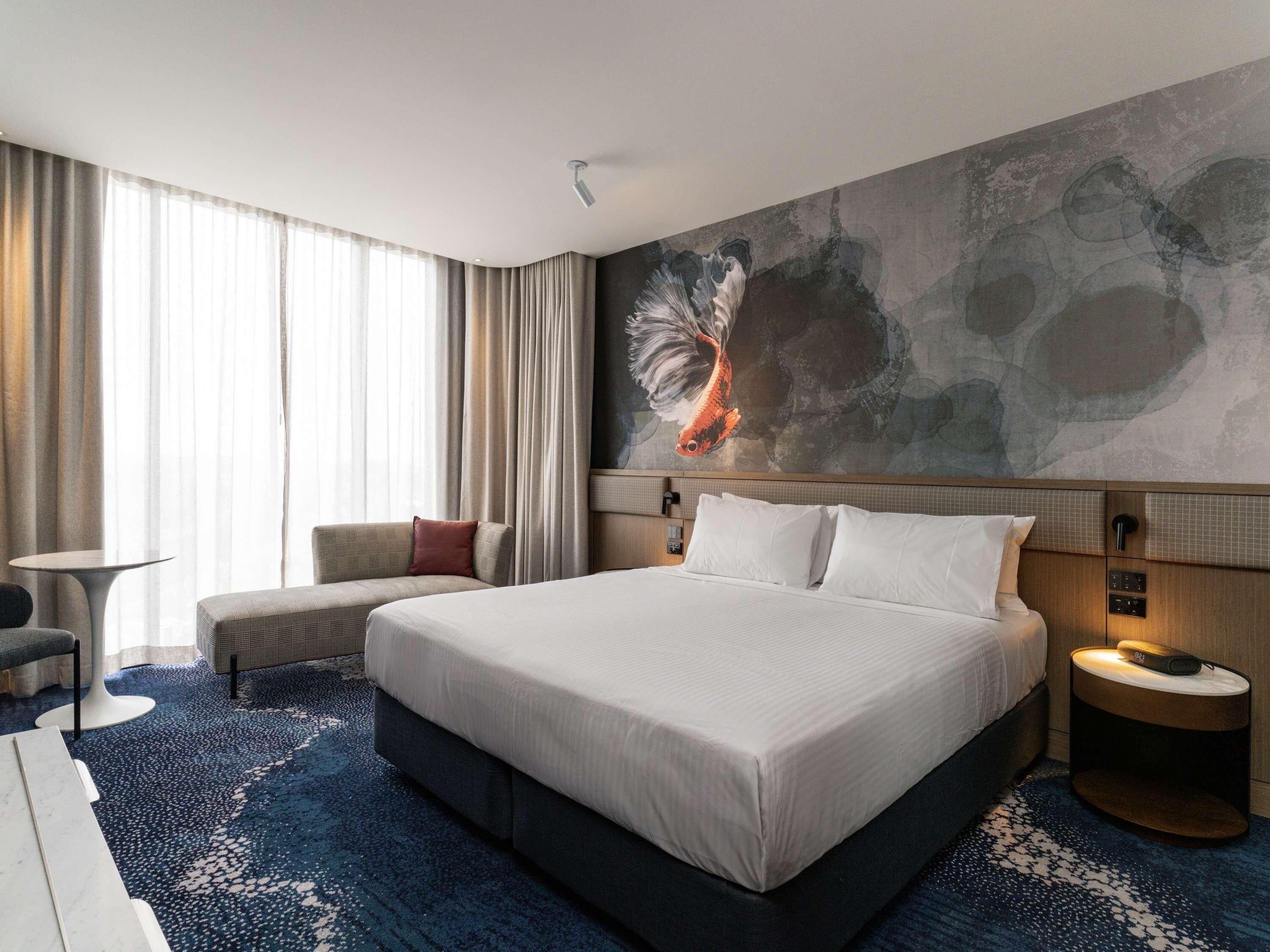 A hotel room with a large bed, a seating area with a chair and ottoman, a small table, and a wall with a large fish painting.