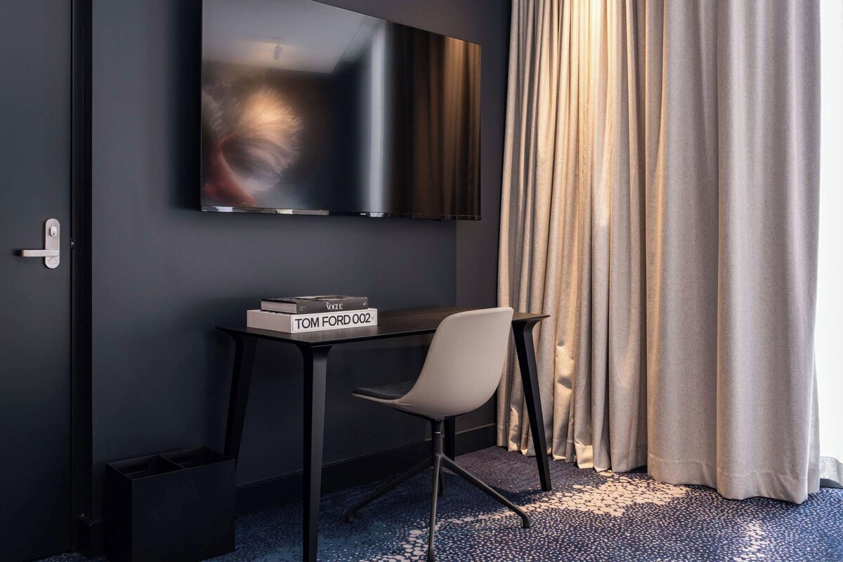 A modern room with a flat-screen TV mounted on the wall, a small table with a chair, and a window with curtains.