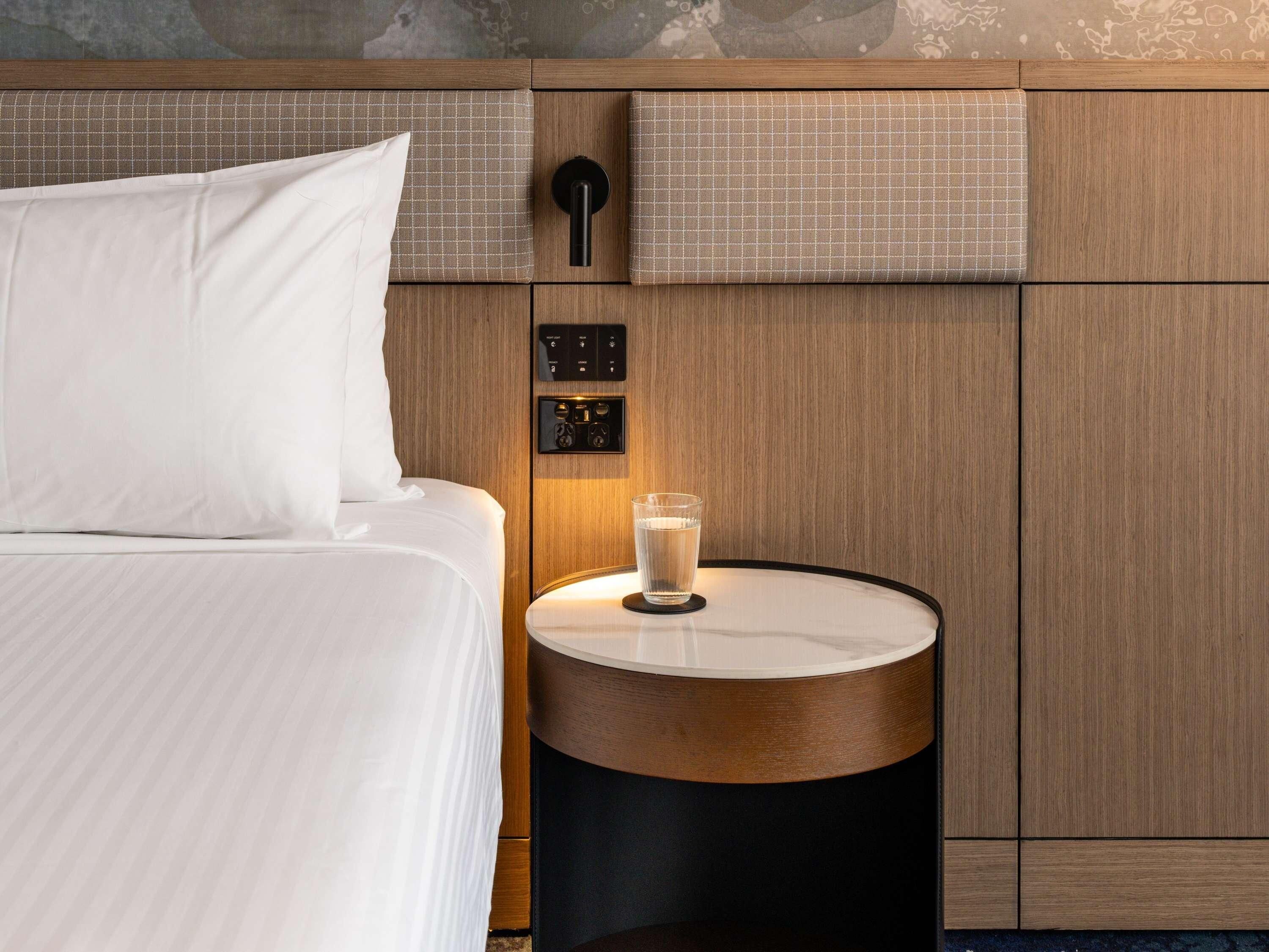 Una habitación de hotel con una cama, una mesita de noche con un vaso de agua, un panel de control y un cabecero de madera.