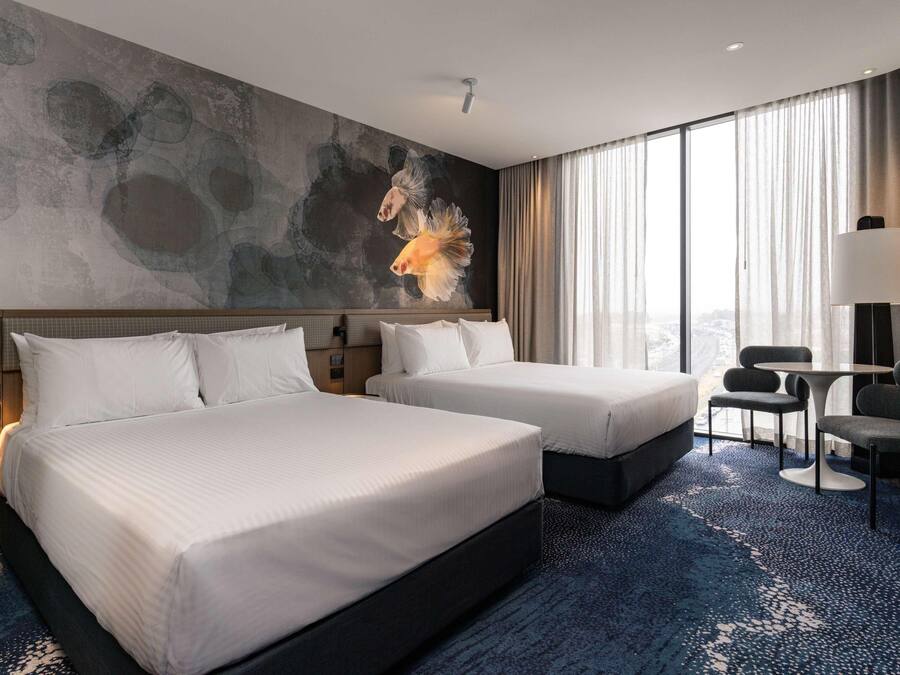Habitación de hotel con dos camas, un ventanal con cortinas, un cuadro en la pared y una pequeña mesa con sillas.