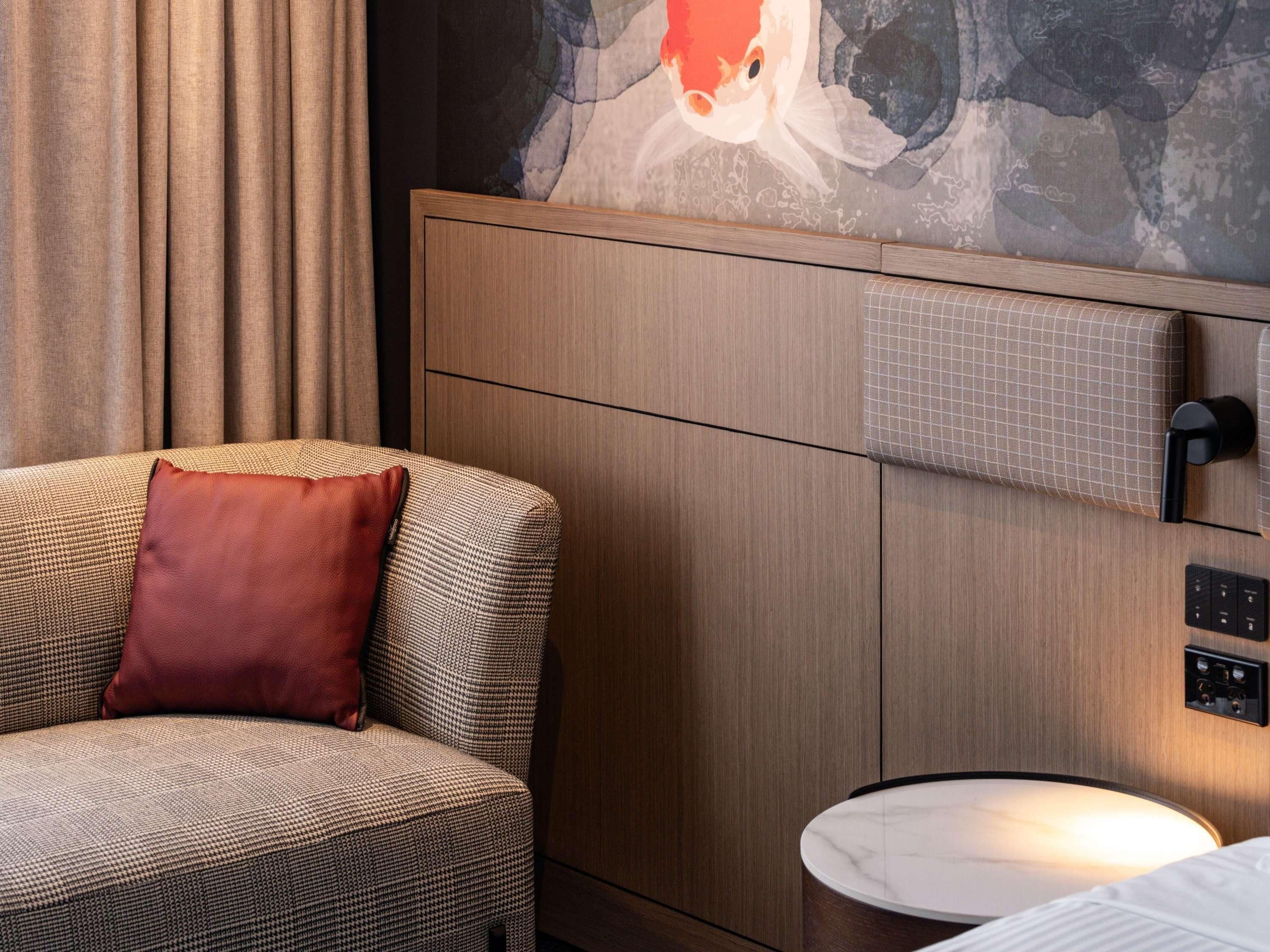 Una habitación de hotel con un sofá a cuadros de piedritas, una almohada roja, una mesita de noche de mármol, un cuadro en la pared y paneles de madera decorativos.