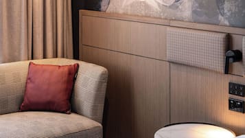 Habitación de hotel con un sofá a cuadros de galgo, una almohada roja, una mesita de noche de mármol, un cuadro en la pared y un panel de control en la pared.