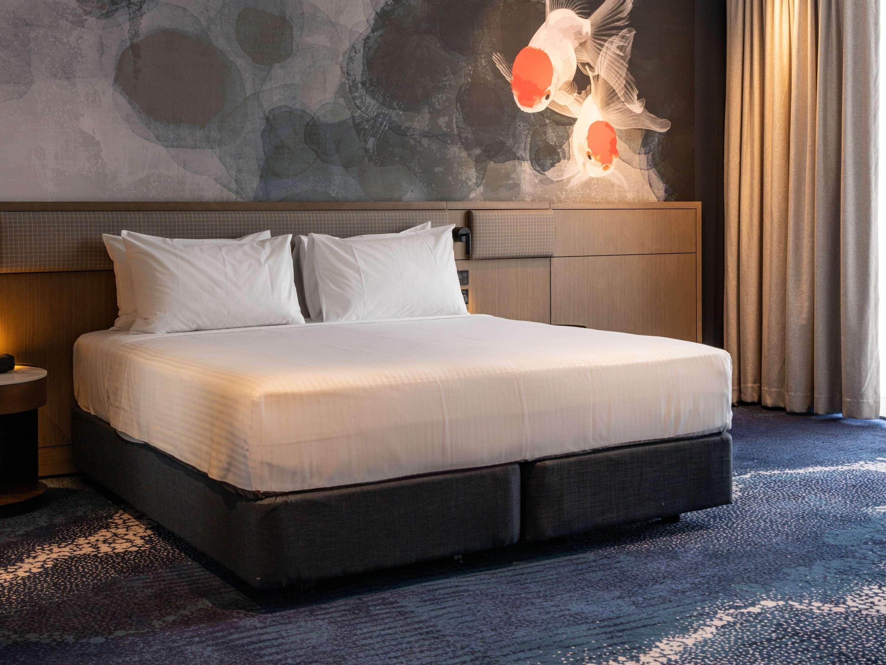 Habitación de hotel con una cama grande, cabecera con diseño texturizado, un cuadro de peces en la pared y una ventana con cortinas.