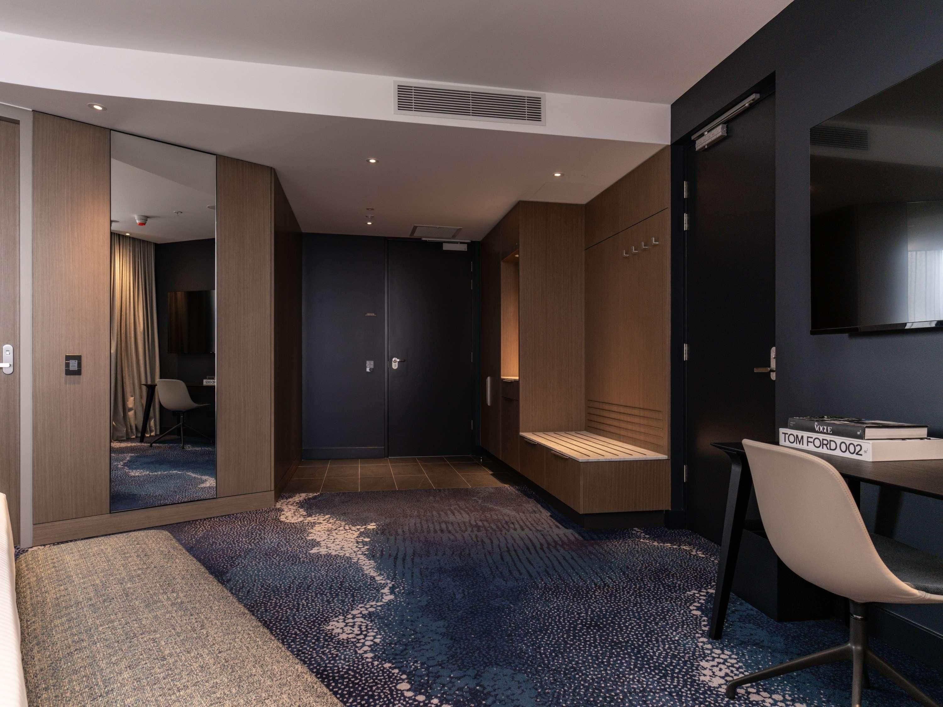 Una habitación de hotel moderna con un espejo grande, un televisor de pantalla plana, una zona de estar con un banco y una pequeña mesa con libros.