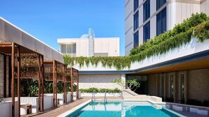 A rooftop swimming pool with a wooden deck, a stone wall, and a building with a modern design in the background. - Novotel Sydney Cabramatta (Sydney)