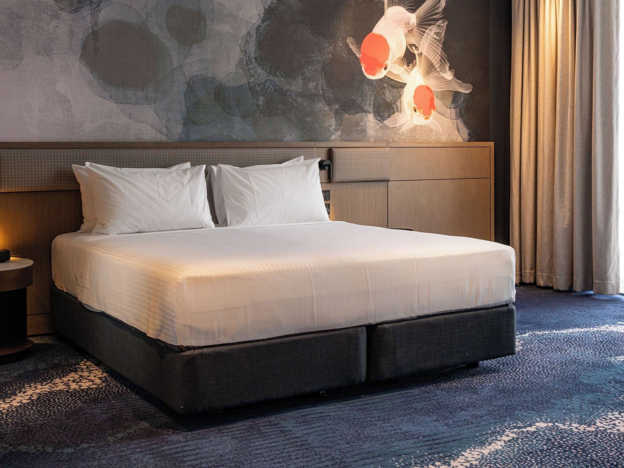Une chambre d’hôtel avec un grand lit, une tête de lit au motif texturé, une fresque murale et une fenêtre avec des rideaux.