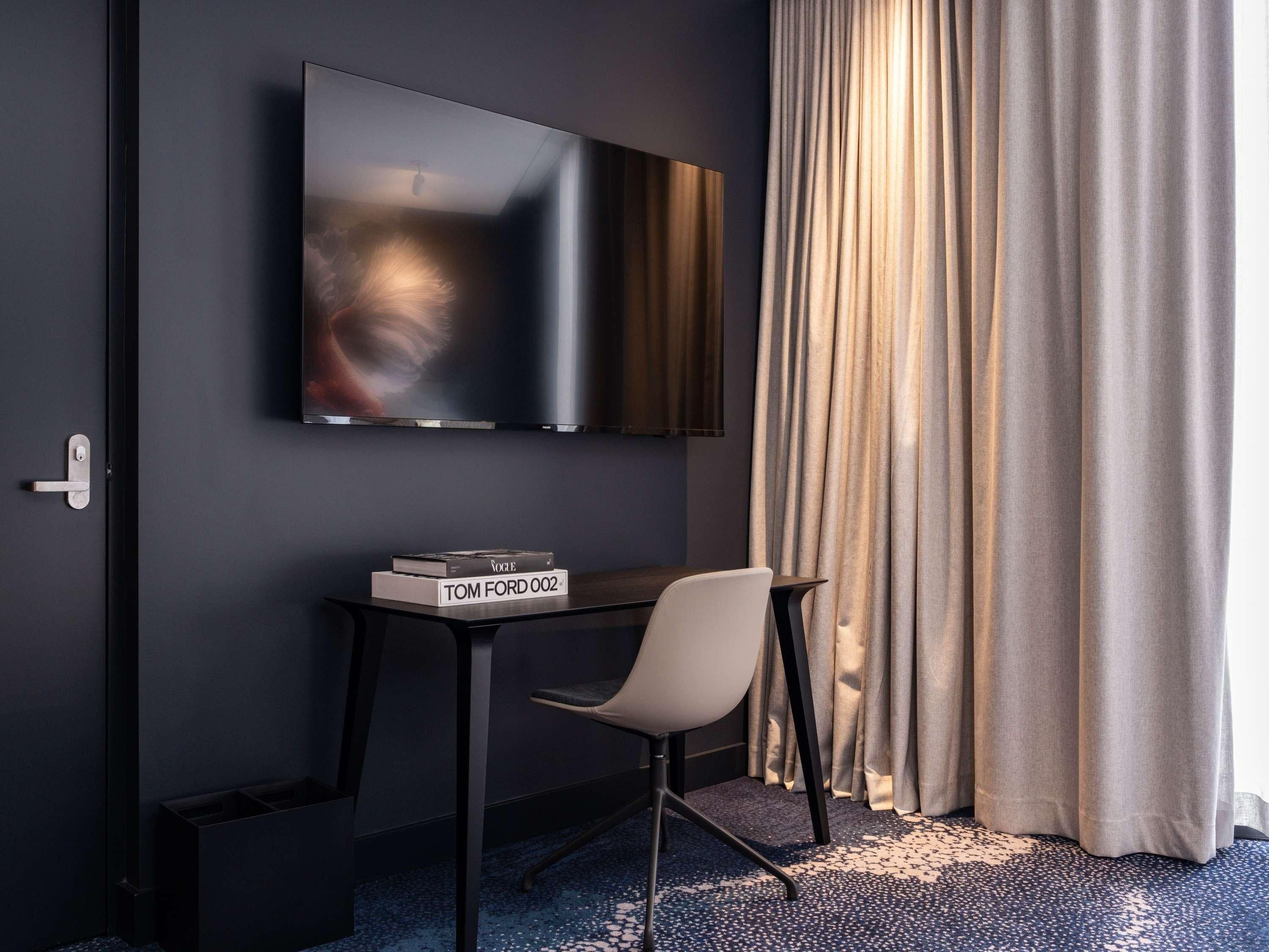 Una habitación de hotel moderna con un televisor de pantalla plana montado en la pared, una mesita con una silla y una ventana con cortinas.