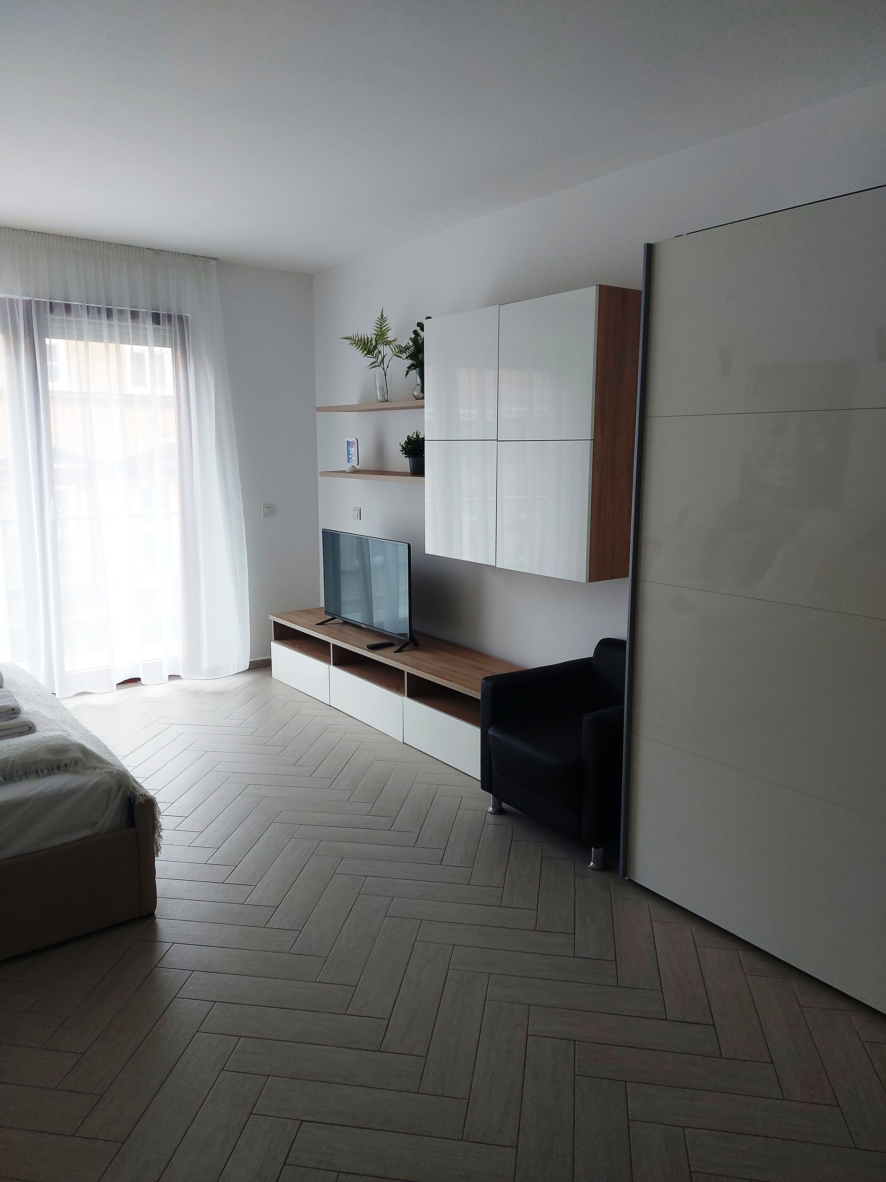 A modern bedroom with a bed, a TV on a wooden shelf, a black chair, and a wardrobe.