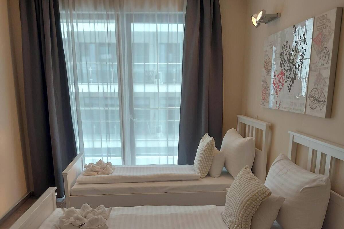A hotel room with two beds, a large window with curtains, a wall-mounted light, and a piece of wall art.