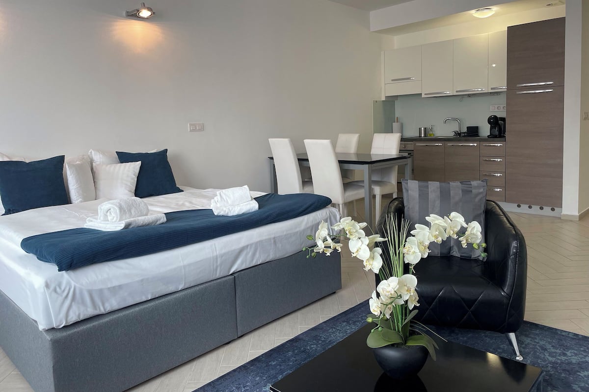 A modern hotel room with a large bed, a dining table, and a kitchen area.