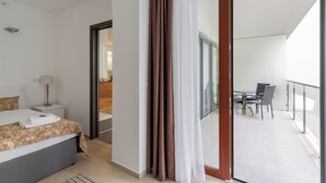 Una habitación de hotel con cama, mesita de noche, lámpara y vistas a un balcón con mobiliario exterior.