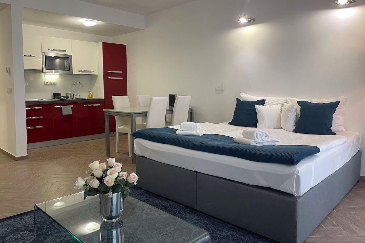A modern hotel room with a bed, bedside table, and a small kitchen area.