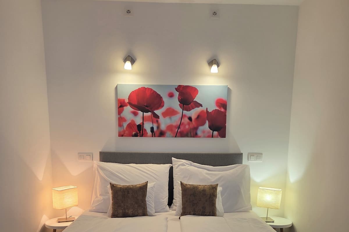 A neatly made bed with white and brown bedding, two bedside lamps, and a large wall art of red flowers.