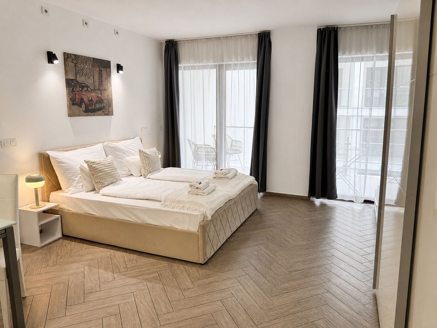 Habitación de hotel moderna con una cama grande, una mesita de noche con lámpara y vistas a la ciudad a través de amplios ventanales.