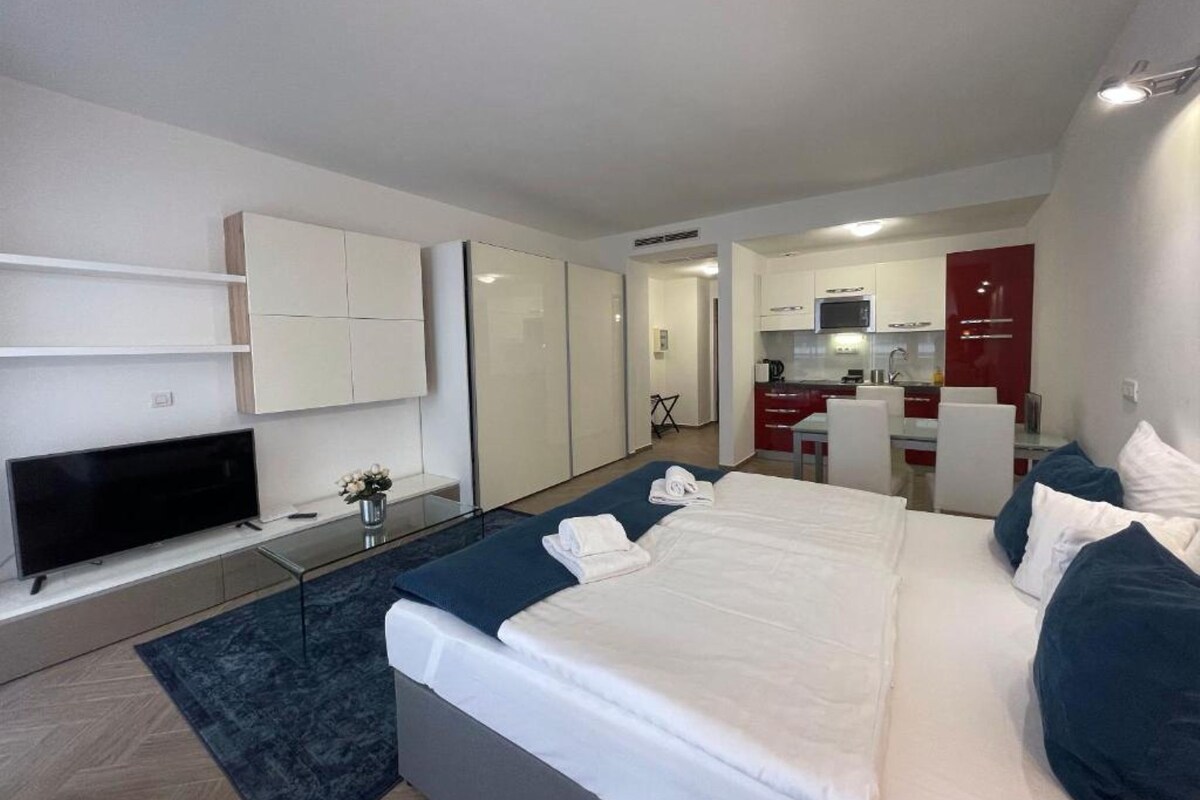A modern hotel room with a large bed, a flat-screen TV, a small desk, and a kitchenette in the background.