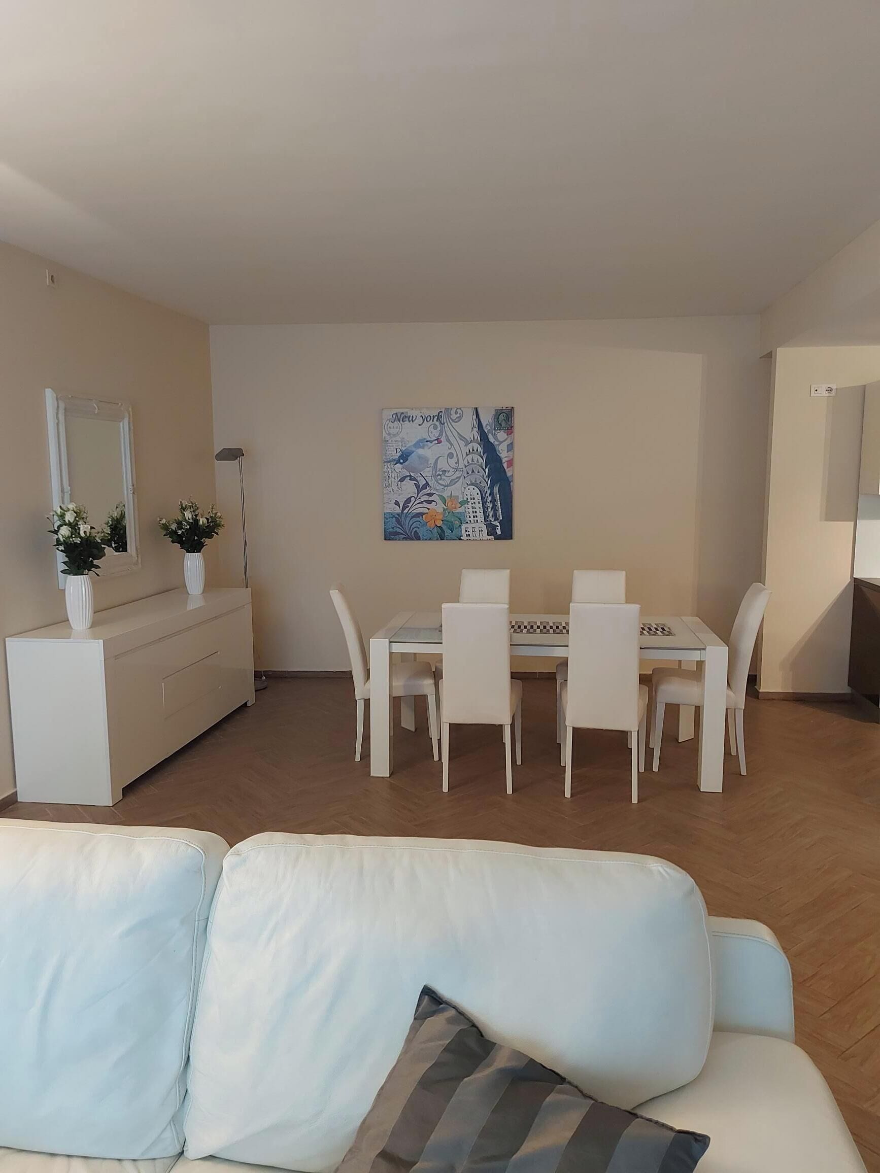 A modern living room with a white sofa, a dining table with chairs, a white cabinet with potted plants, and a painting on the wall.