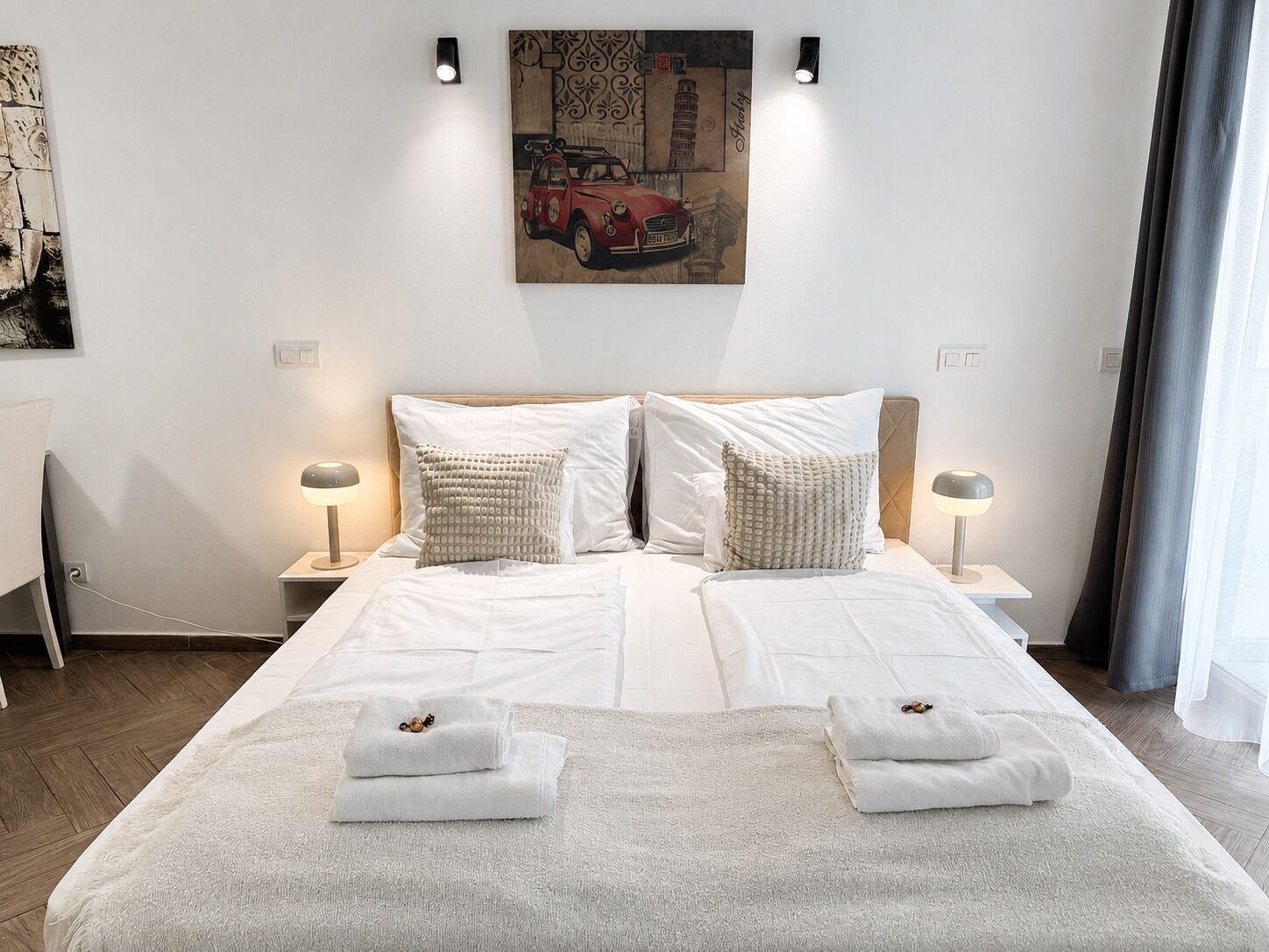 A hotel room with a bed, two bedside lamps, a painting on the wall, and a window with curtains.