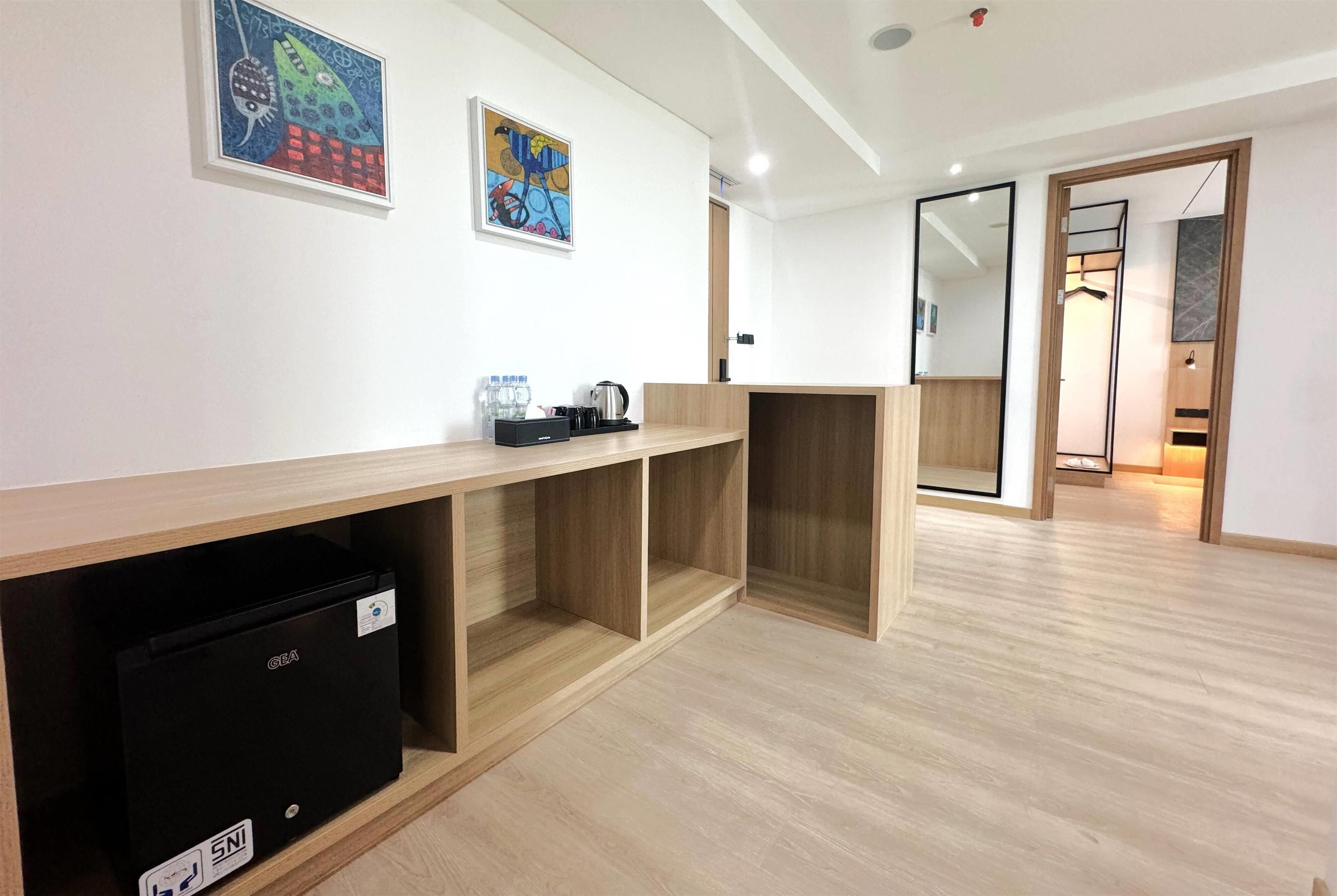 A hotel room with a wooden floor, a desk with a kettle, a black storage unit, and two framed artworks on the wall.