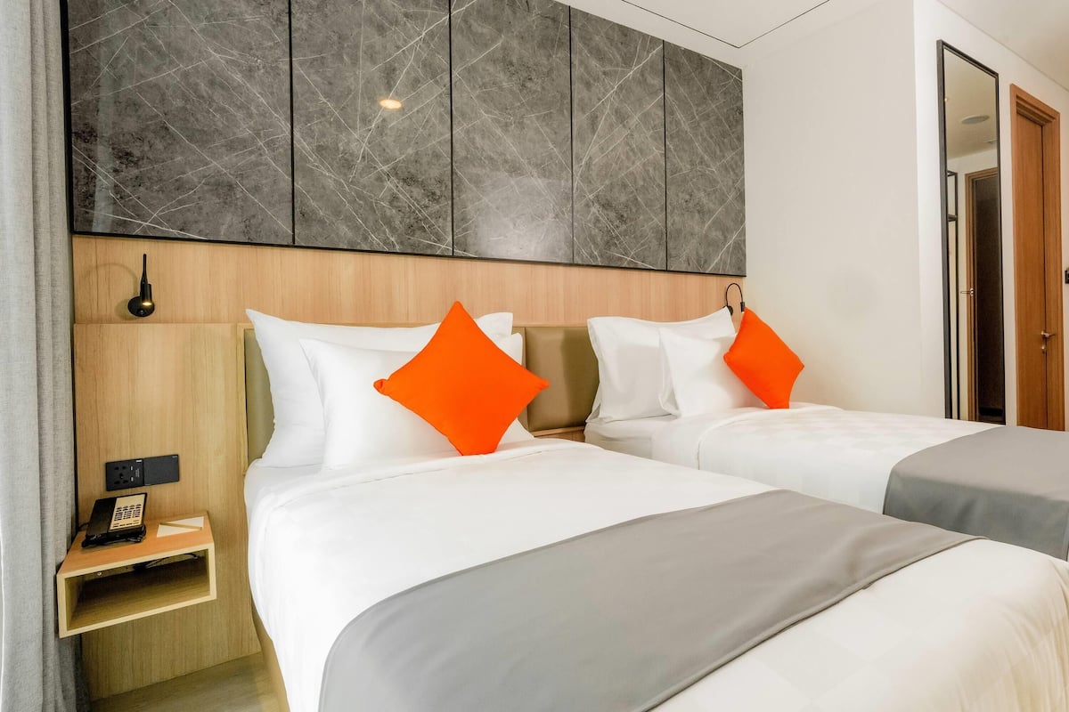 Habitación de hotel con dos camas, cada una con ropa de cama blanca y almohadas naranjas. Se observa un cabecero de madera, una mesita de noche con un teléfono y una lámpara de pared.