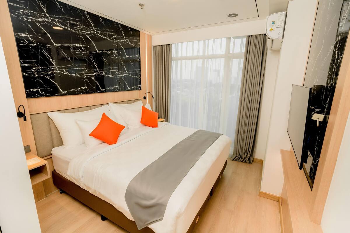 Una habitación de hotel con una cama grande, pared de mármol, televisor y ventana con cortinas.