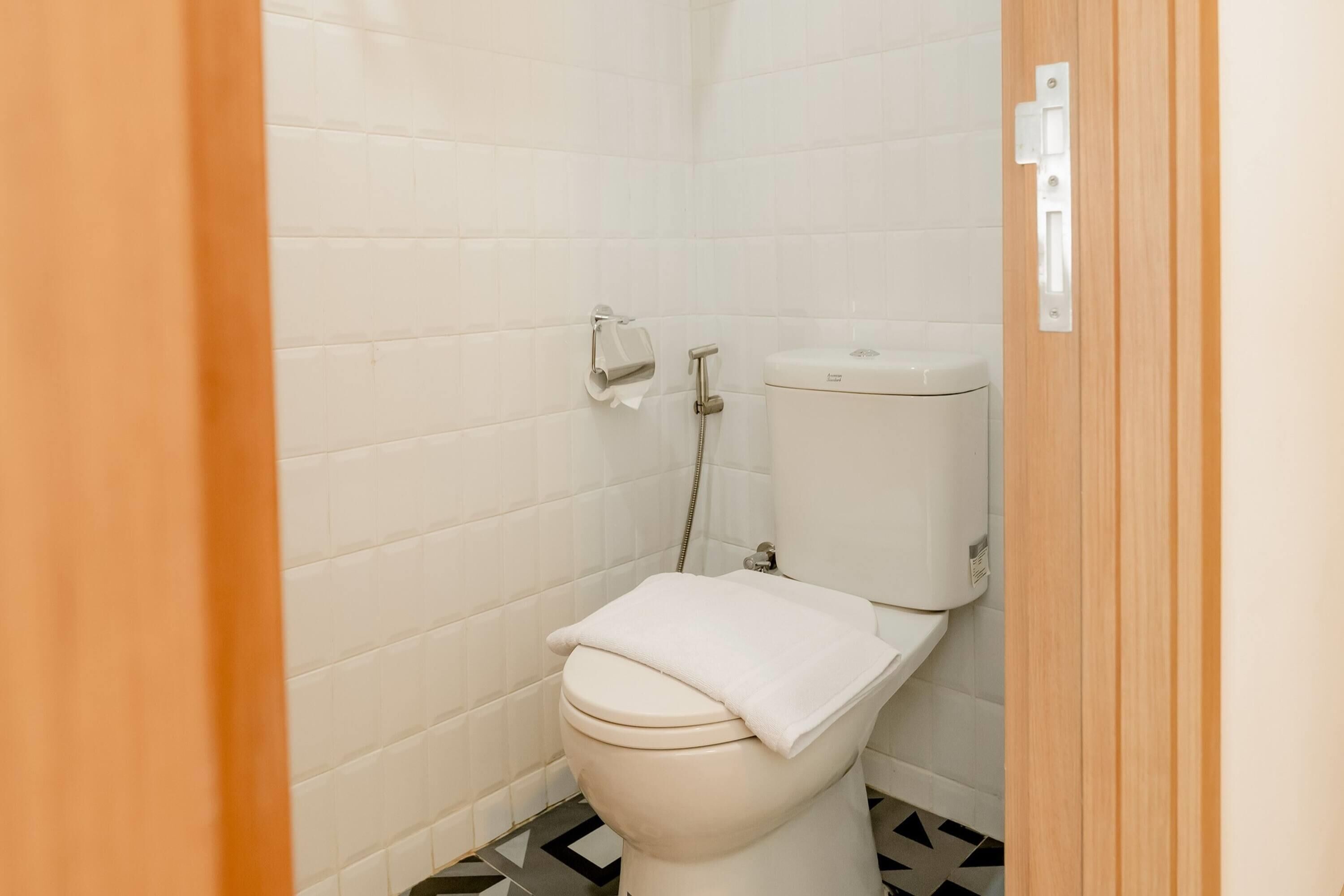 A small, clean bathroom with white tiles, a toilet, a paper towel dispenser, and a showerhead.