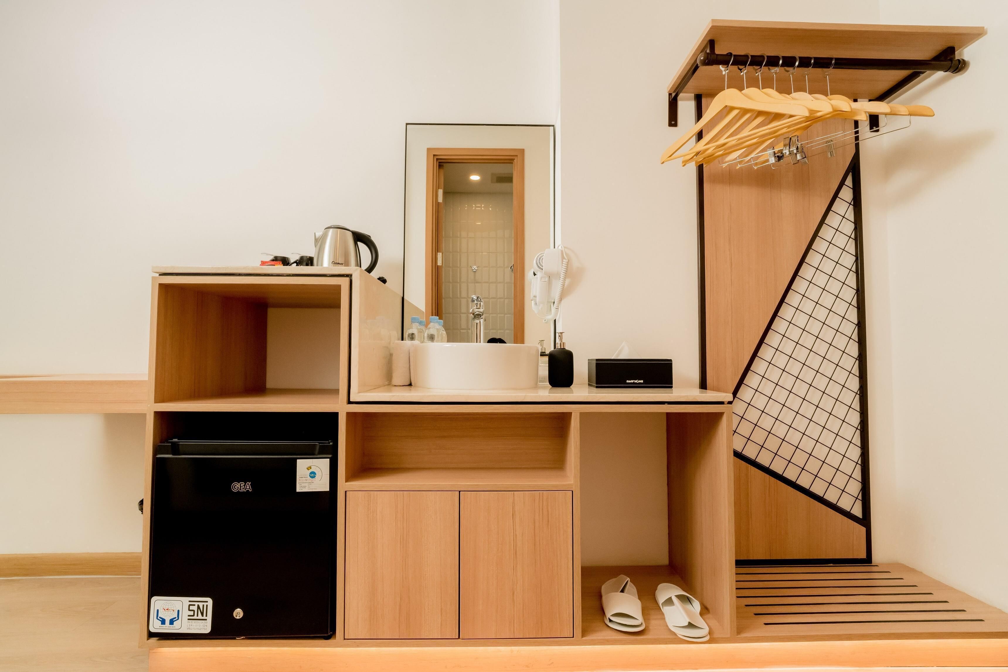 A compact hotel room with a wooden cabinet, a sink, a mirror, a drying rack, a heater, and a small refrigerator.