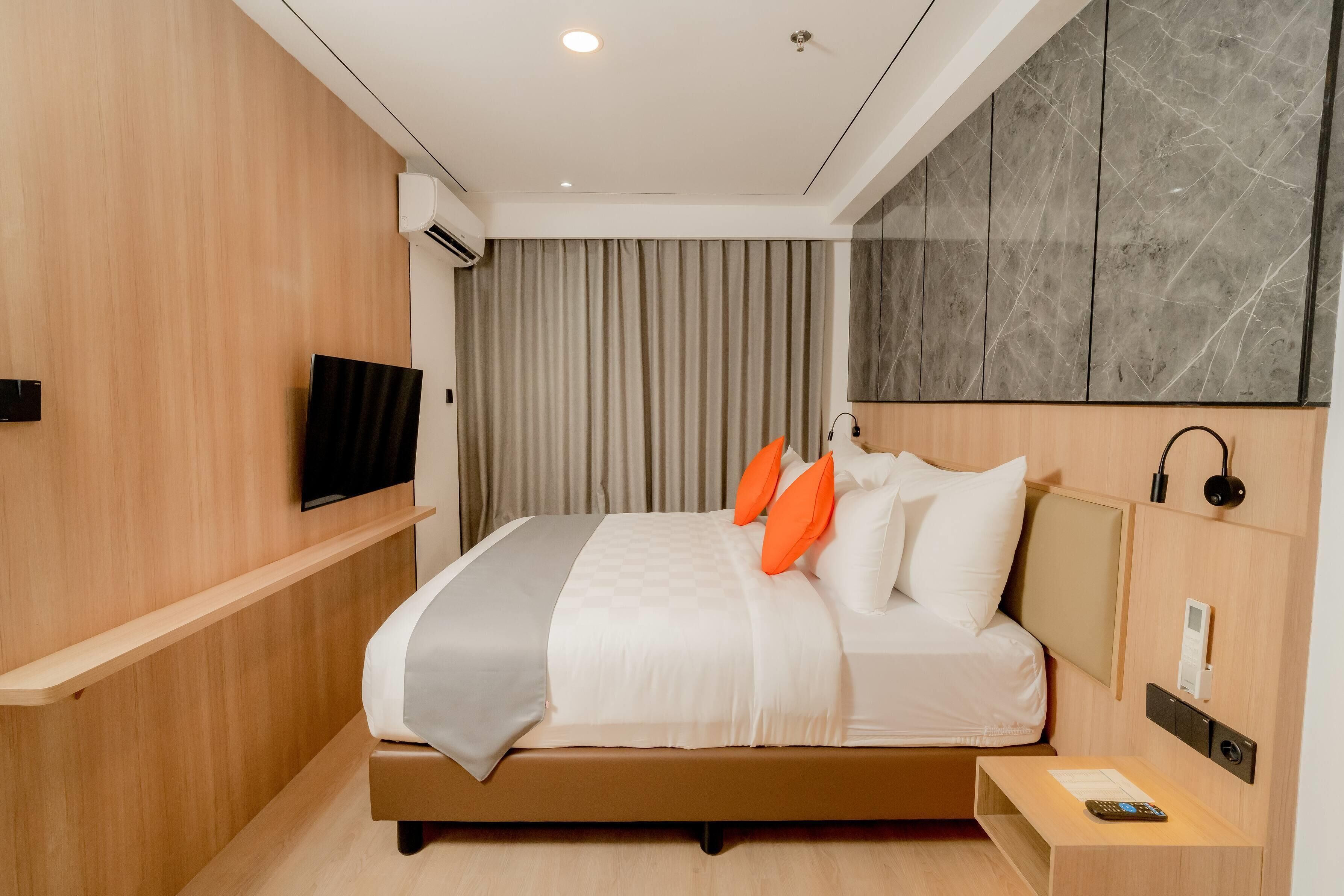 Habitación de hotel moderna con pared de madera, televisor de pantalla plana, cama con ropa blanca y almohadas anaranjadas, mesita de noche con un control remoto y una pared decorativa de mármol.