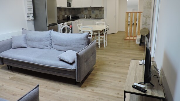 A modern living room with a grey sofa, a dining table with chairs, a kitchen area with white cabinets, a washing machine, and a TV on a wooden stand. - townhouse (saujon)