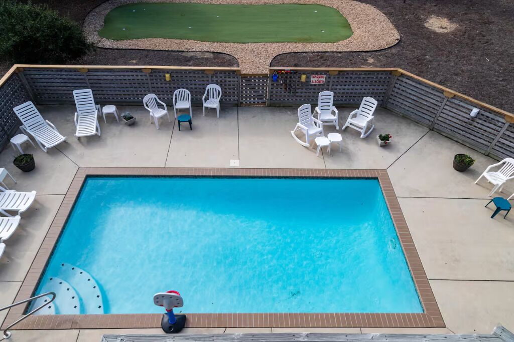 A rectangular swimming pool with a surrounding deck, white lounge chairs, and a small garden area with a green lawn and a putting green.