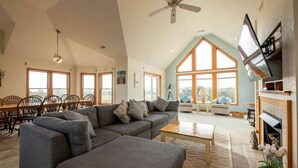 Un salón espacioso con un sofá modular grande de color gris, una mesa de centro de madera y una chimenea. Varias ventanas ofrecen luz natural y vistas al exterior.