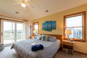 A bedroom with a large bed, a ceiling fan, a window with a view, and a nightstand with a lamp and books. - Sandy Pants-Walk to Beach! (Duck)