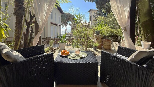 An outdoor patio with wicker furniture, a table with fruits and a cup, and a view of buildings and trees. - Apartment Niko Private Garden Wifi  (Opatija)