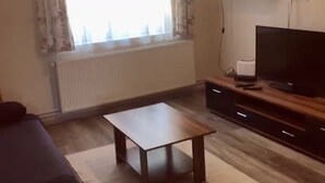 Una sala de estar con sofá, una mesa de centro, un televisor sobre un mueble de madera, un radiador y una ventana con cortinas.