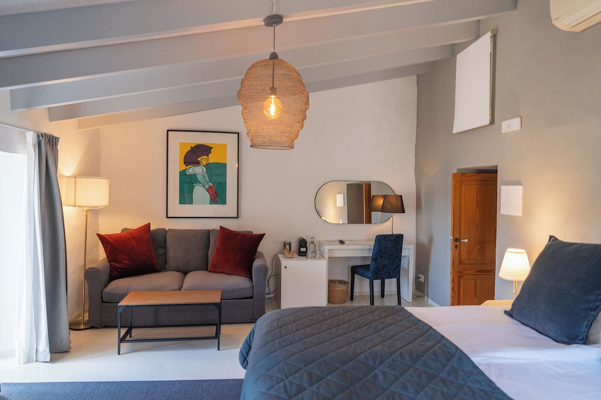 A modern hotel room with a bed, a desk, a chair, a sofa, and a hanging light fixture.