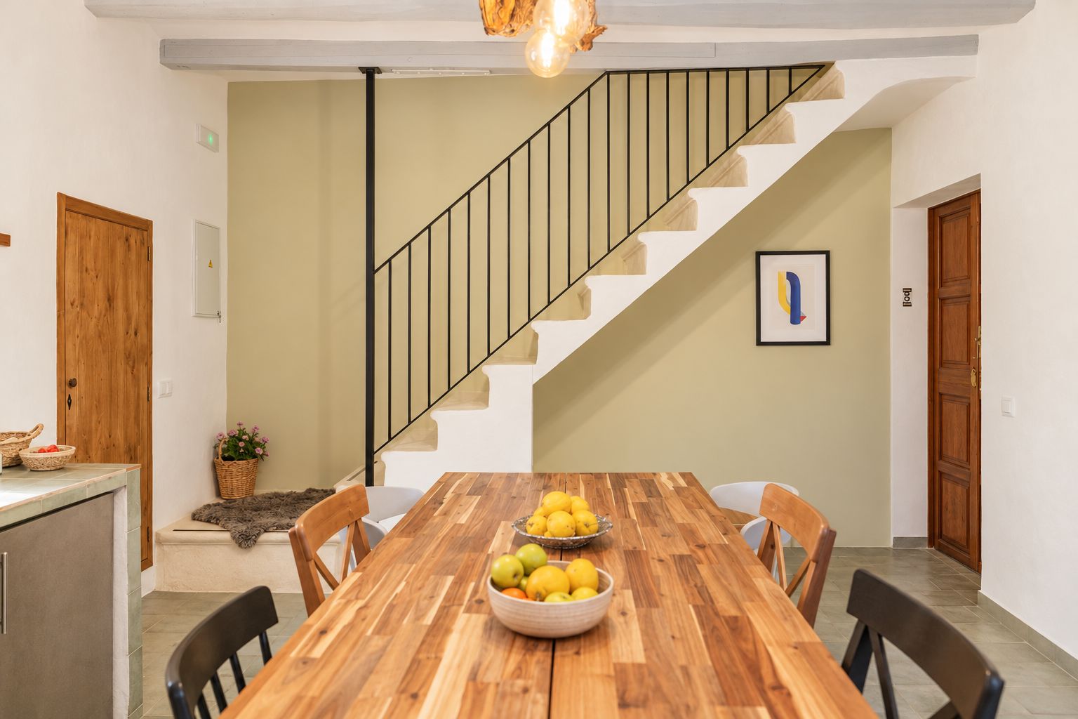 A dining area with a wooden table, chairs, and a bowl of fruit. A staircase with a black railing is visible in the background.
