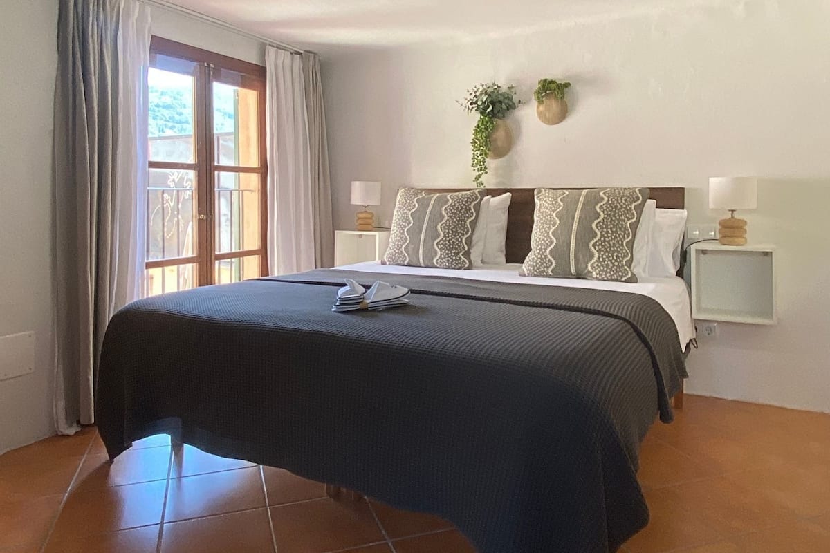 A bedroom with a bed, two bedside tables, a window with curtains, and two wall-mounted plants.