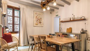A dining area with a wooden table, chairs, and a fruit bowl. There is a window with curtains, a wall-mounted artwork, and a shelf with glasses and bottles. - Solleric Petit Hotel (Sóller)