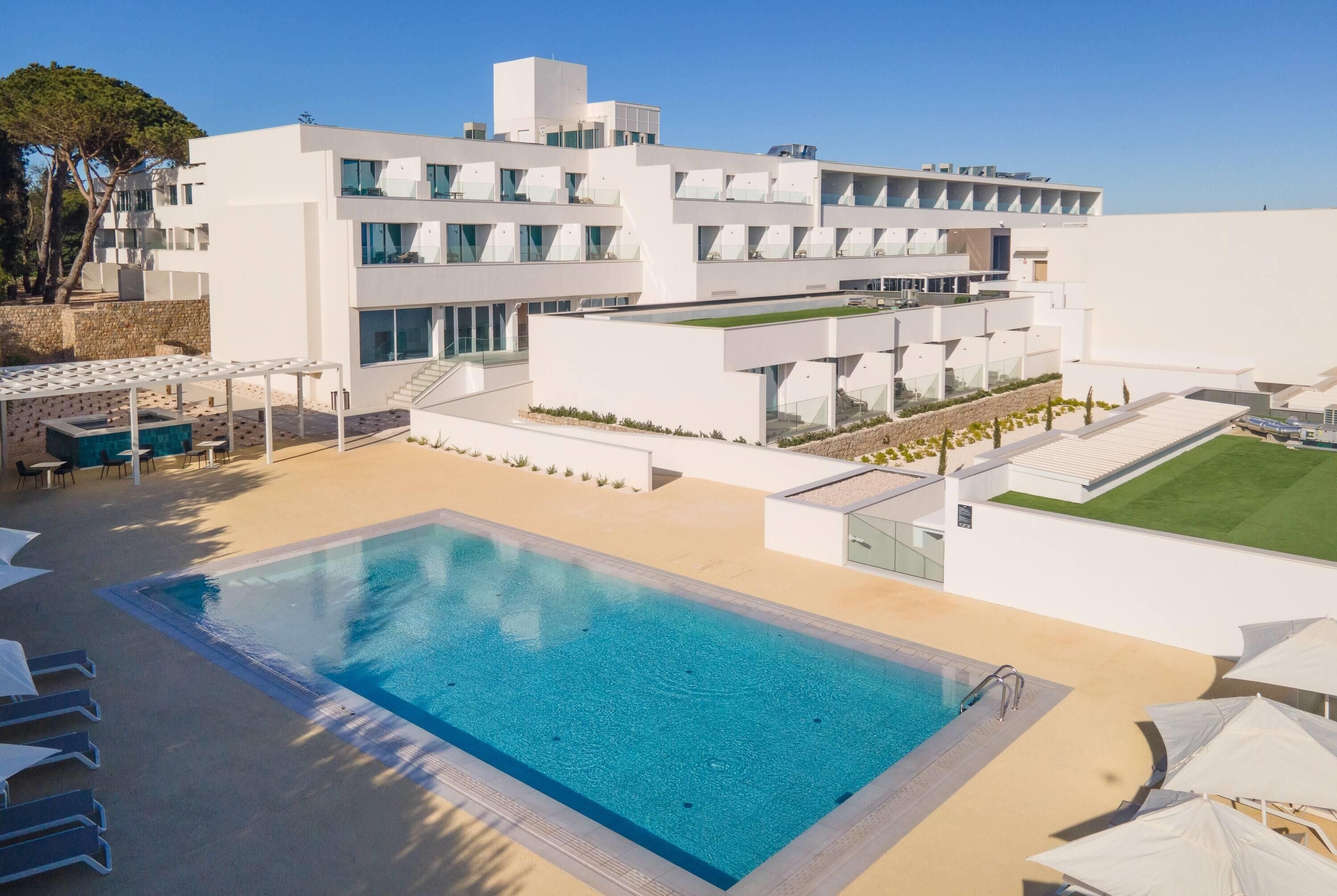Un hotel moderno con piscina, mobiliario exterior y un césped cuidado.
